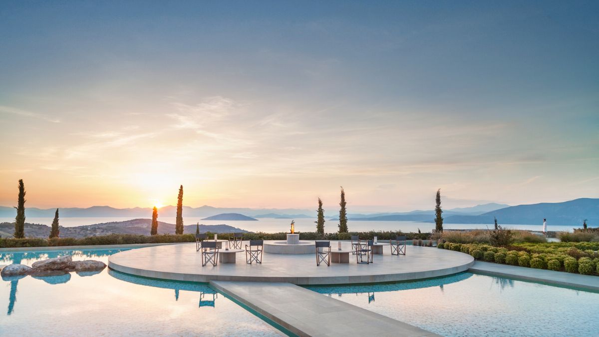 A serene outdoor space featuring a circular seating area with chairs and a central fire pit, overlooking a tranquil pool. The scene is framed by lush greenery and hills in the background, with a vibrant sunset casting warm colors across the sky.