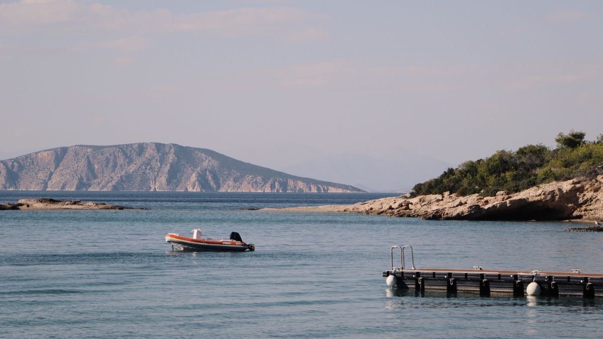 A small boat floats in calm waters near a dock, with a mountainous island in the background under a clear sky.