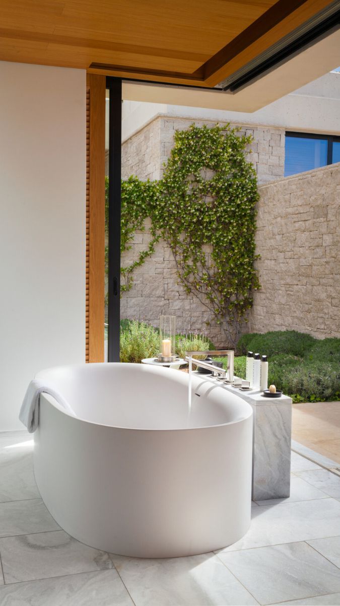 A modern, freestanding white bathtub sits elegantly in a spacious bathroom. Natural light floods in through large sliding glass doors that open to a serene outdoor space with a stone wall and lush greenery. Minimalist decor and accessories enhance the tranquil atmosphere.