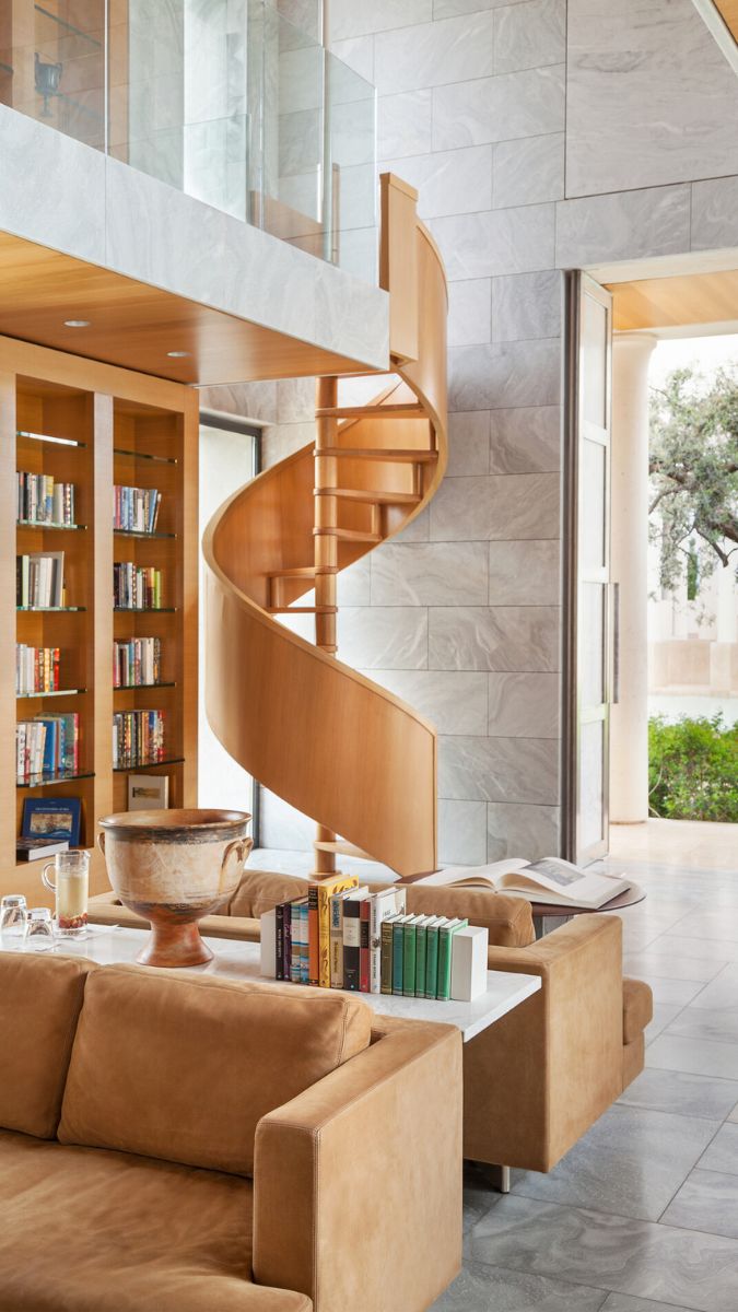 A modern living room features a spiral wooden staircase, sleek stone walls, and built-in bookshelves filled with books. A cozy seating area with brown sofas is visible, along with a coffee table and decorative items. Large windows allow natural light and greenery from outside.