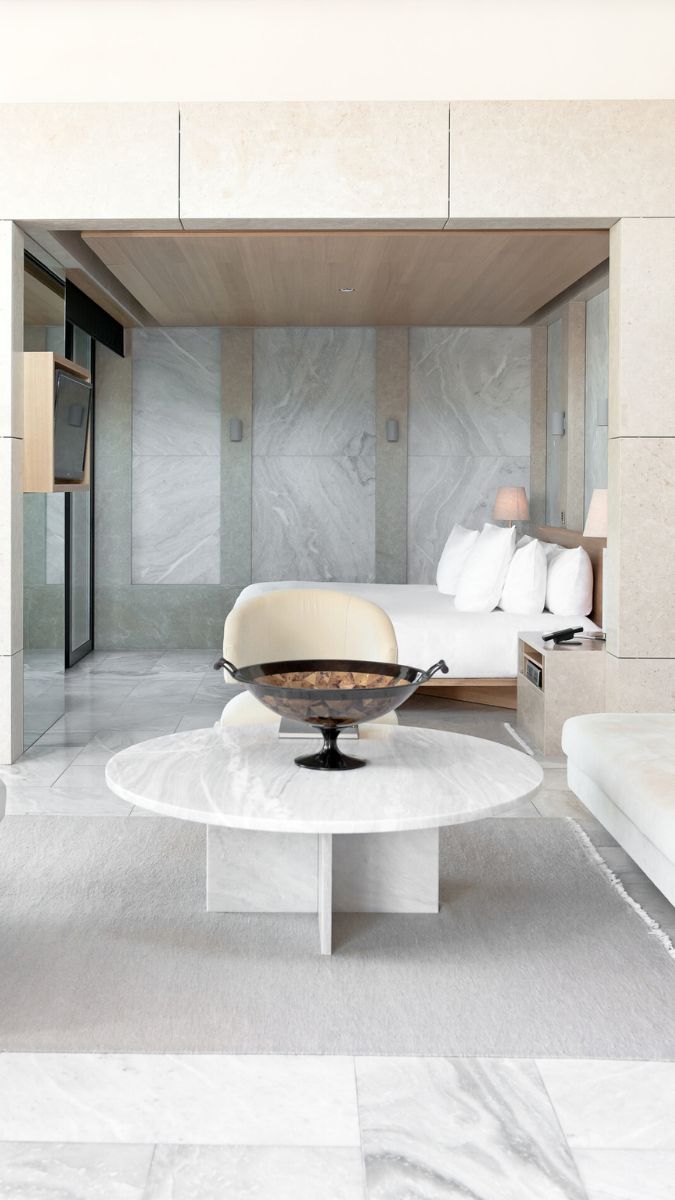 A modern, upscale hotel suite featuring a spacious layout with marble flooring, a plush bed with white linens, and elegant furniture. A round marble coffee table sits in the center, adorned with a decorative bowl. Natural light floods the room from large windows, enhancing the luxurious atmosphere.