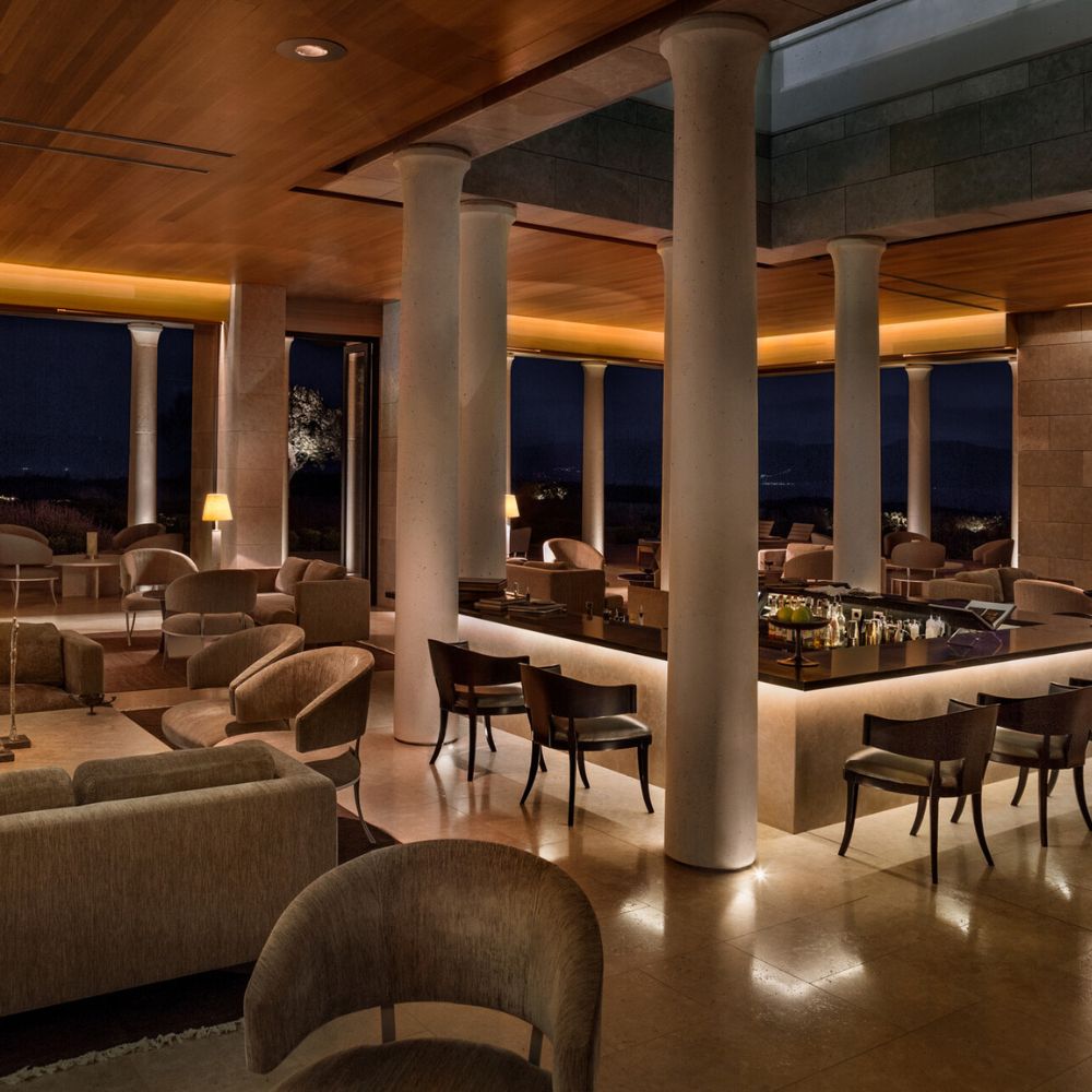 A stylish lounge area with modern furnishings, featuring soft lighting, tall columns, and a bar counter. The space exudes a warm, inviting atmosphere, set against a night backdrop with expansive windows showing a distant view.