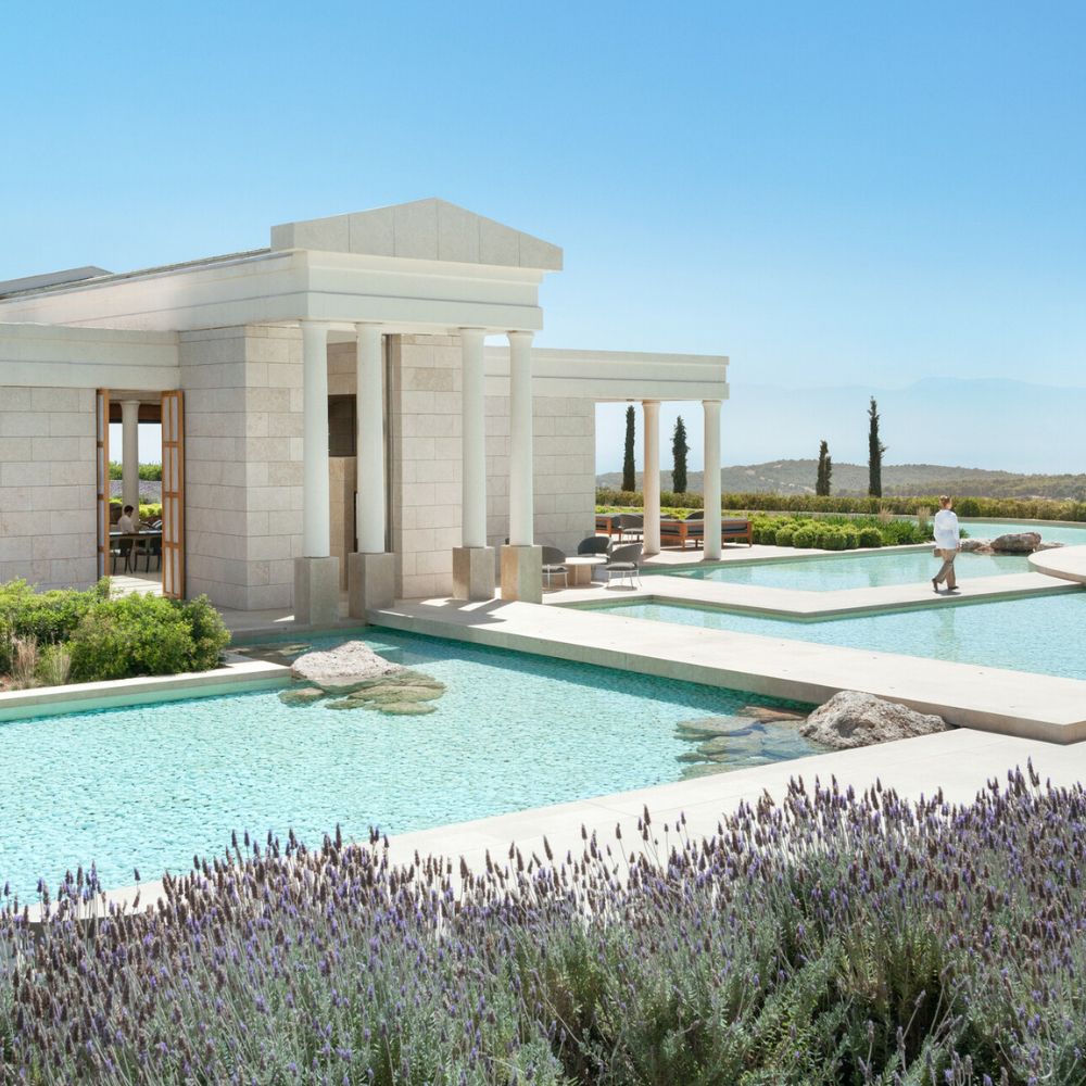 A modern architectural building with classical columns surrounded by serene pools and lush greenery. Lavender flowers are in the foreground, and distant hills are visible under a clear blue sky. A person walks along the path beside the water.