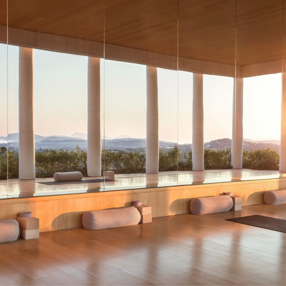 A serene yoga studio with large floor-to-ceiling windows showcasing a sunrise over rolling hills. The wooden floor features soft bolsters neatly arranged, and the warm sunlight creates a peaceful ambiance.