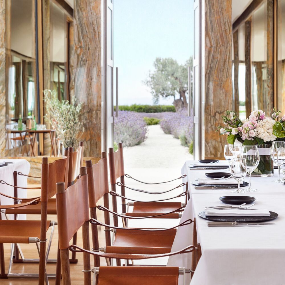 A beautifully arranged dining area with elegant tables set for a meal. Leather-strapped chairs surround crisp white tablecloths, and large windows frame a scenic view of lavender fields and olive trees outside. Fresh flowers serve as a centerpiece.