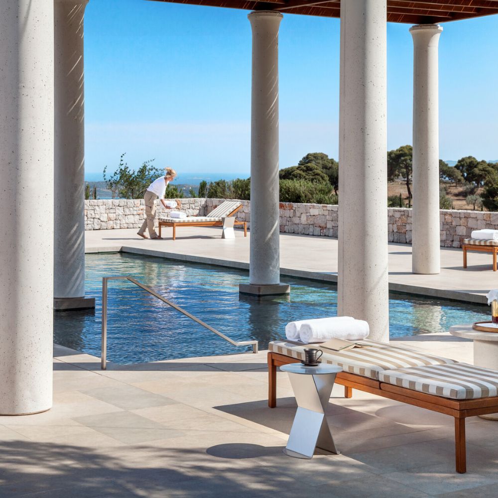 A serene outdoor area featuring a swimming pool with a Reflection edge, surrounded by columns and sun loungers. A person in light clothing is seen arranging towels near the pool, with a clear blue sky and distant trees in the background.