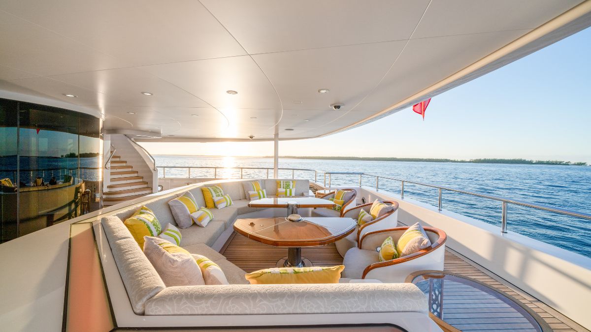 A spacious yacht deck features a comfortable seating area with plush cushions and a round wooden table, overlooking the serene water at sunset. The scene is enhanced by a warm glow and the silhouette of distant land.