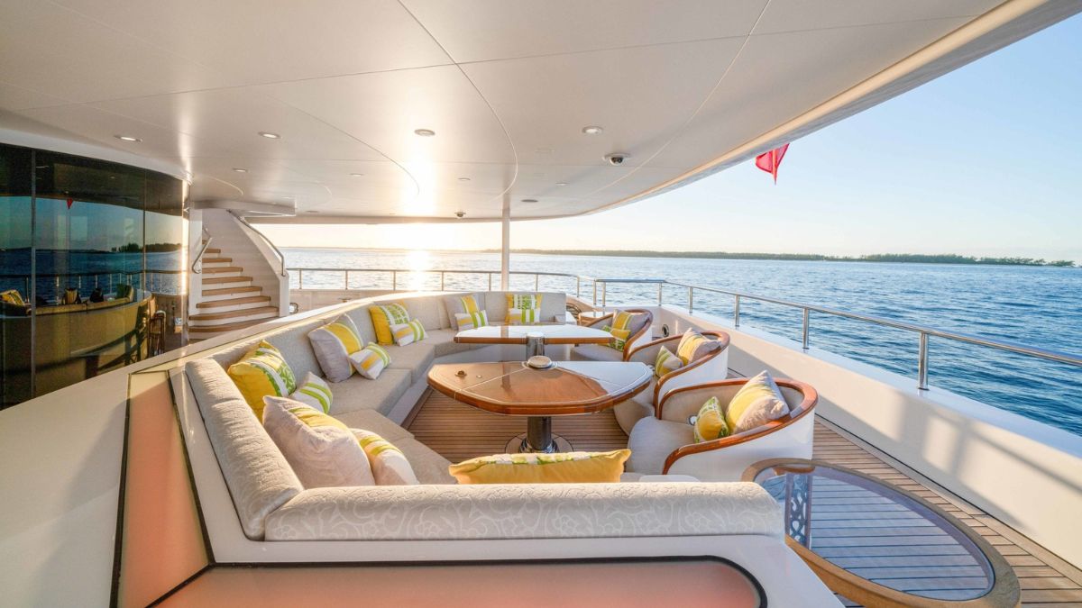 Luxurious outdoor lounge area on a yacht, featuring a circular table surrounded by plush seating with colorful cushions. A serene view of calm waters and a sunset in the background.