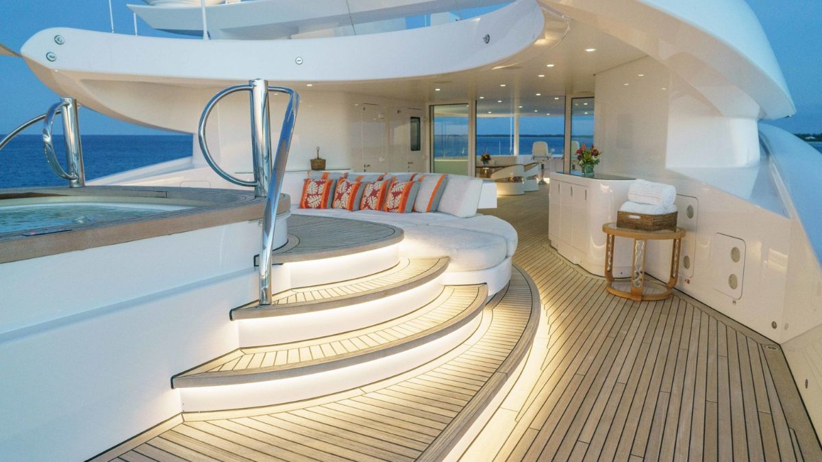 A luxurious yacht deck features elegant seating with orange and white pillows, inviting ambient lighting, and steps leading to a hot tub, all overlooking a serene ocean at dusk.