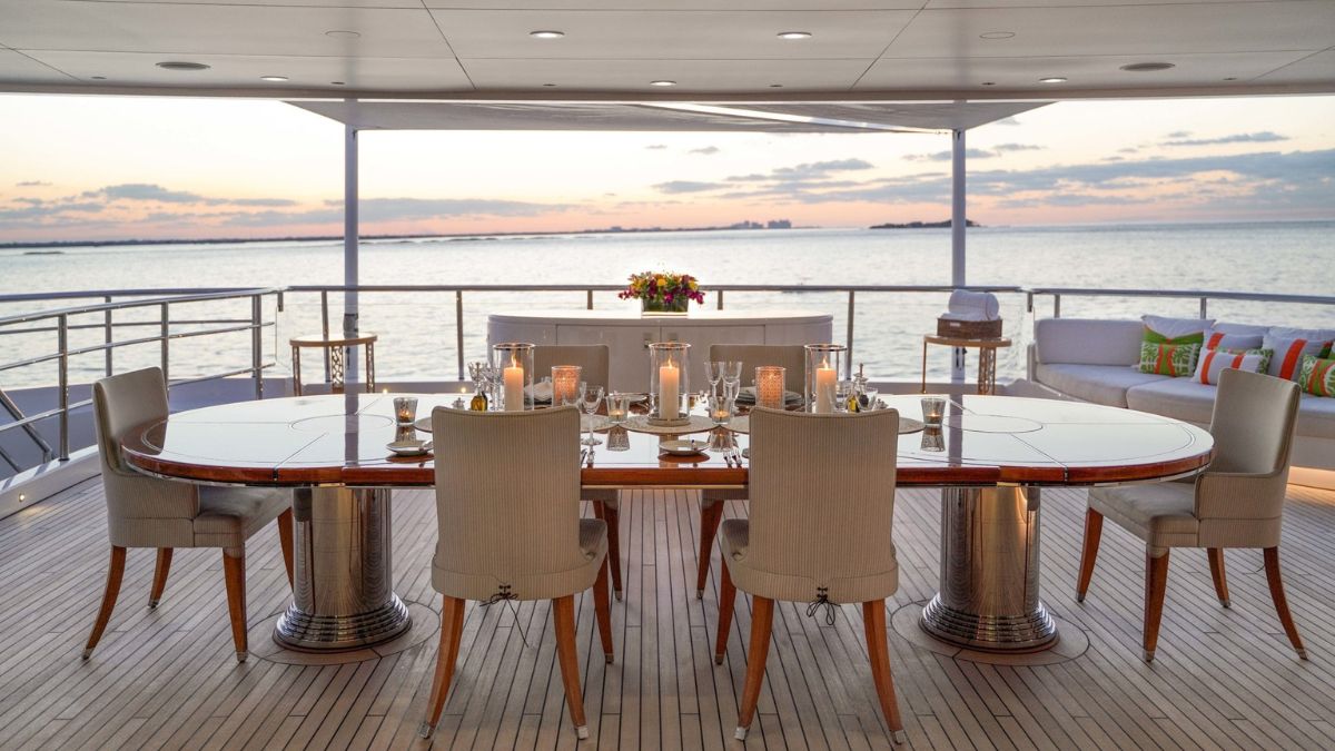 A beautifully set dining table on a yacht, surrounded by elegant chairs, with candles and floral arrangements. The sunset casts a warm glow over the tranquil water in the background.