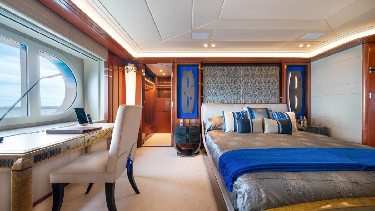 Luxurious bedroom featuring a large bed with blue and gold linens, an elegant desk with a chair, and a window showcasing a scenic view. The decor includes intricate wood paneling and stylish lighting.