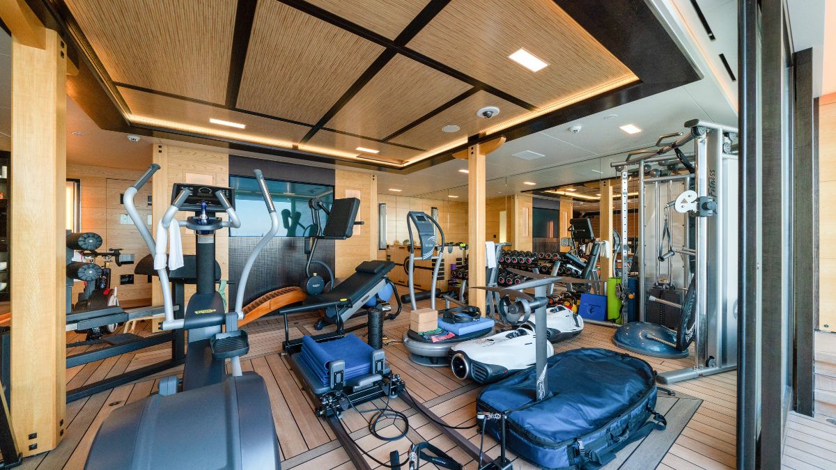 A well-equipped gym onboard a yacht, featuring various exercise machines, weights, and fitness equipment. The space has a modern design with wooden accents and large windows.