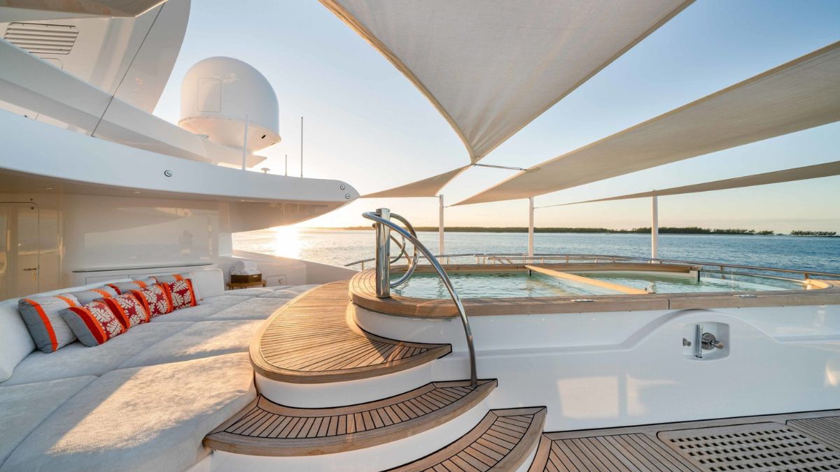 A luxurious yacht deck features plush seating with orange and white pillows, a circular jacuzzi, and elegant wooden stairs. The sun sets over calm waters, creating a serene atmosphere.