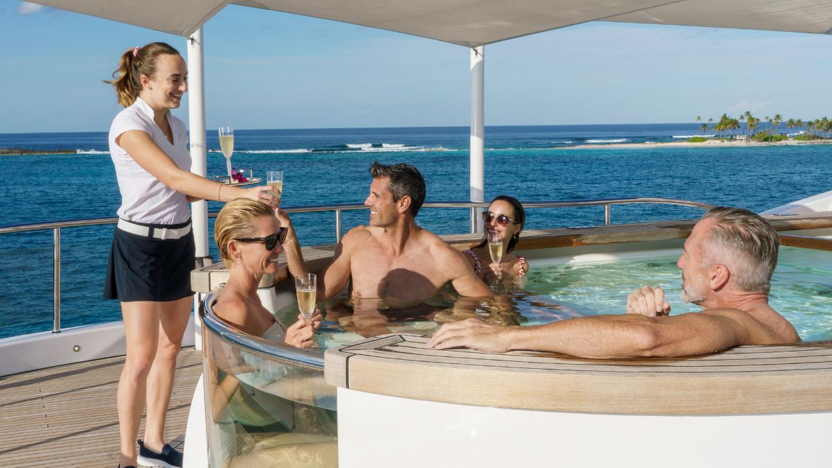 A group of four people enjoying drinks in a hot tub on a yacht, with a waitress serving champagne. The ocean and a tropical island are visible in the background.