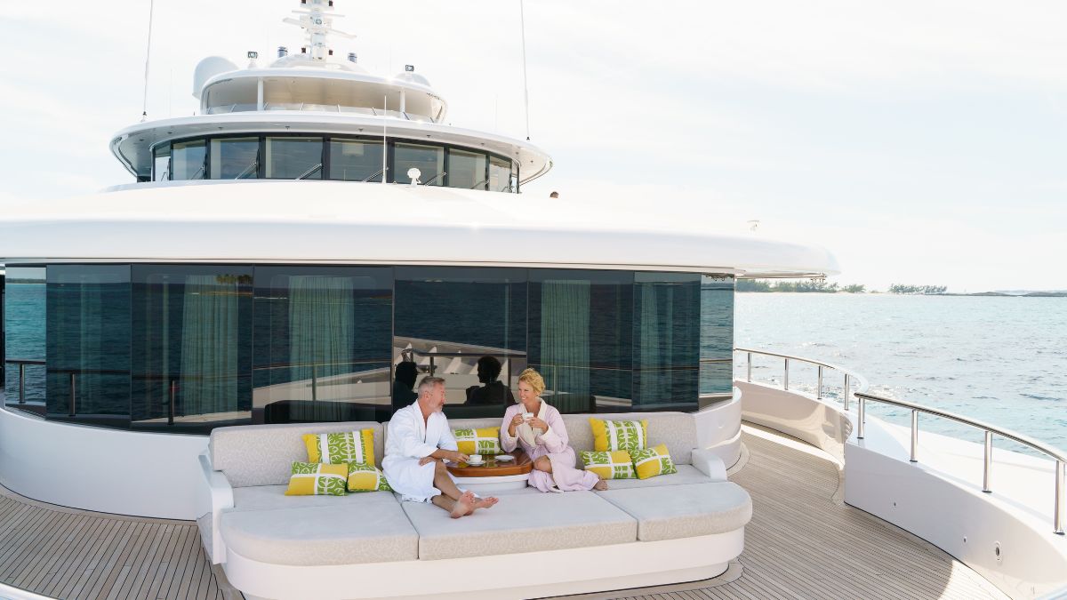 A man and a woman relax on a spacious yacht deck, seated on a large sofa adorned with colorful cushions. They are enjoying drinks and engaging in conversation, with a calm ocean and horizon in the background.