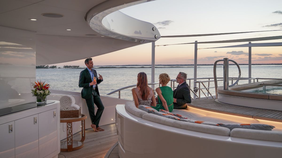 A group of four people enjoys a sunset view on a yacht's deck. One man stands in front, engaged in conversation with the others, who are seated. The scene features a modern, luxurious setting with water in the background.