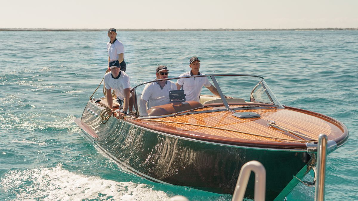 A wooden boat glides through clear turquoise water with four people onboard. Two are seated in the front, while two stand at the rear, all wearing white shirts and caps. The sun shines brightly, highlighting the boat's polished wood finish and the serene surroundings.