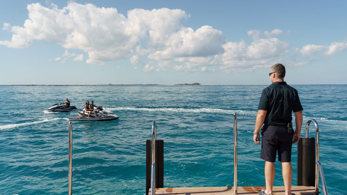 A man stands on a dock observing two jet skis gliding across a turquoise sea under a bright blue sky with fluffy clouds.