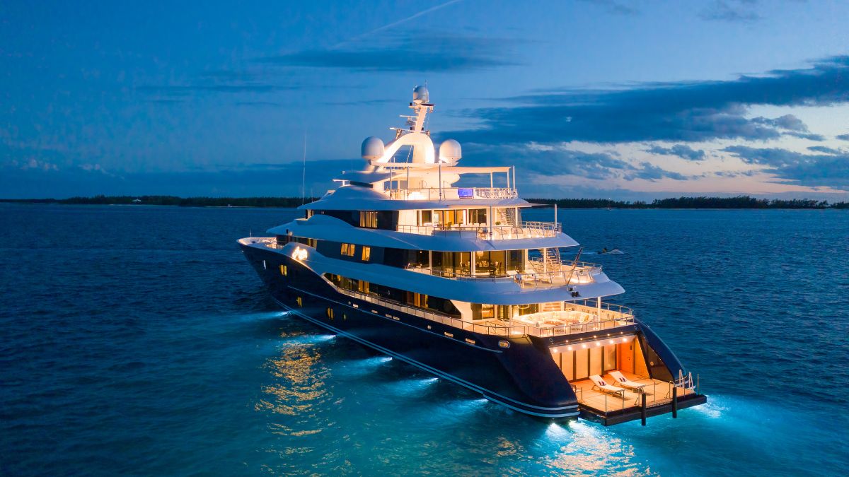 A luxurious yacht illuminated at sunset, gliding on calm waters with a sleek design and multiple decks. The sky transitions from blue to orange as the sun sets on the horizon.