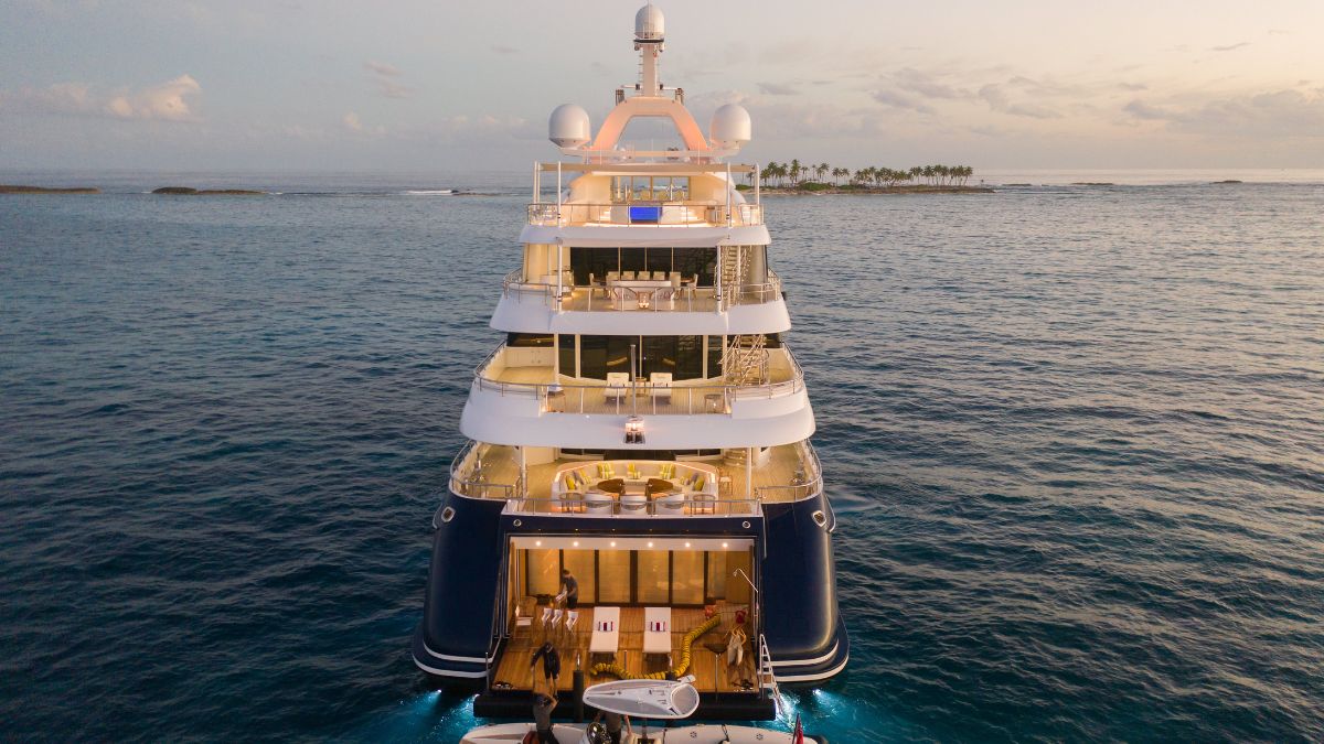 A luxurious multi-deck yacht anchored in calm waters during sunset, showcasing elegant furnishings and outdoor spaces. Palm trees and an island are visible in the background.