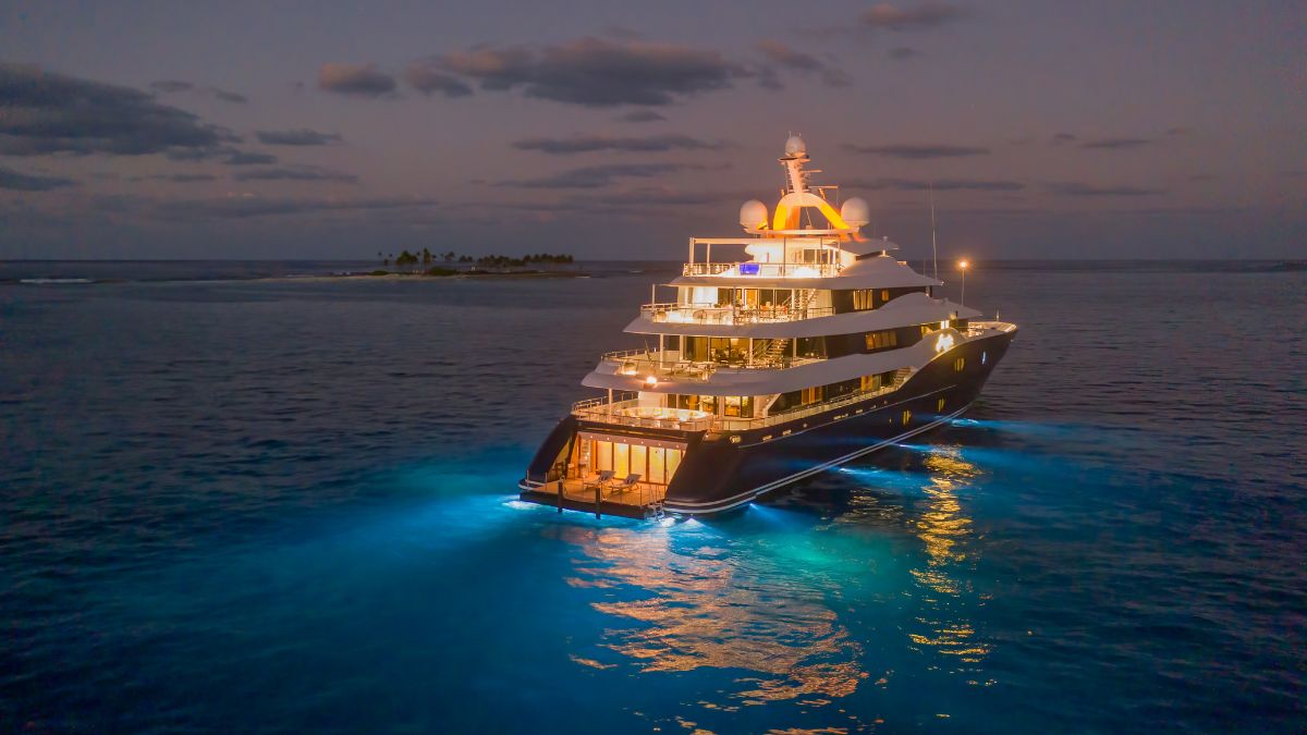 A luxurious yacht illuminated at night, anchored in tranquil waters with a glowing blue underlight. A serene island silhouette is visible in the background under a cloudy sky.