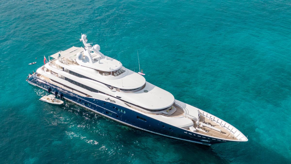 A large luxury yacht anchored in vibrant turquoise waters, featuring multiple decks and sleek design elements. A smaller boat is alongside, highlighting its size and opulence.