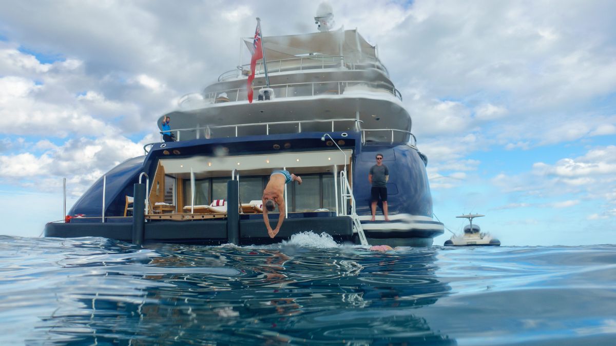 A person diving off the back of a luxury yacht into clear blue water, with another person standing on the yacht deck and a smaller boat nearby. The sky is partly cloudy, creating a serene atmosphere.