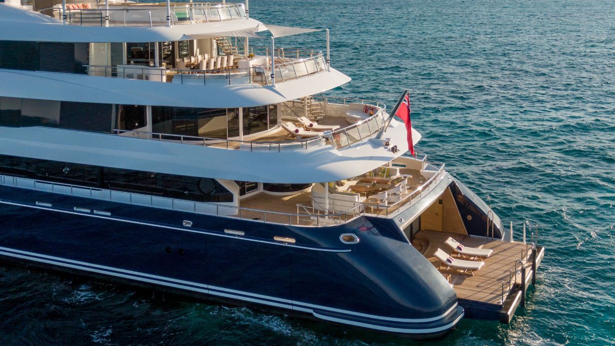 A luxurious yacht anchored on calm blue waters, showcasing multiple levels with sun loungers and elegant seating areas. The yacht features a sleek design with a white and navy colour scheme, and the flag is visible on the bow.