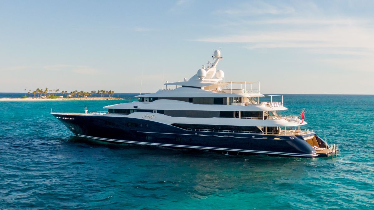 A large, luxurious yacht anchored in vibrant turquoise waters, with a small tropical island and palm trees in the background under a clear blue sky.