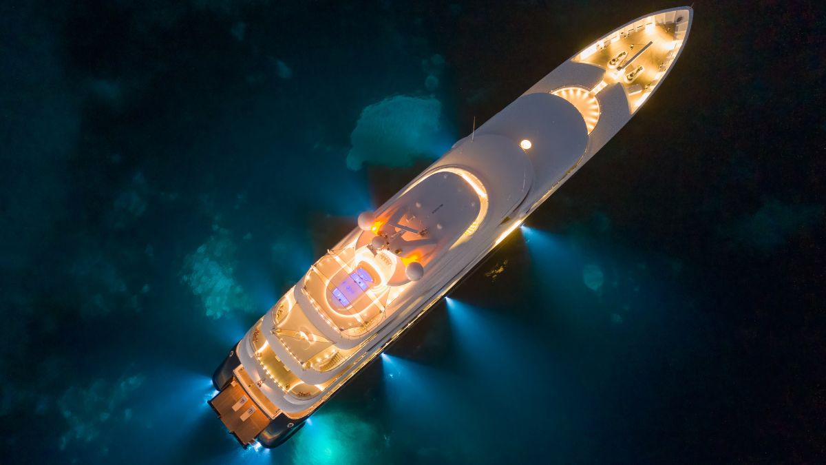A luxurious yacht illuminated with warm lights, anchored in clear, dark waters. The deck features a spacious sunbathing area and an inviting pool, with vibrant underwater lighting highlighting the surrounding seascape.