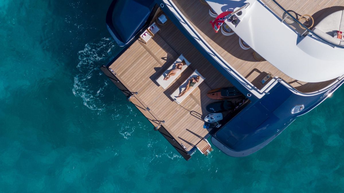 Aerial view of a luxury yacht with two sunbathers lounging on the deck over turquoise waters.