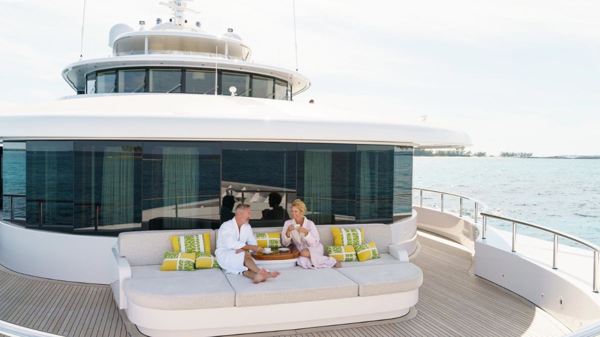 A couple relaxes on a yacht's spacious deck, enjoying drinks while dressed in robes. Colorful cushions decorate the seating area, with a serene ocean view in the background.