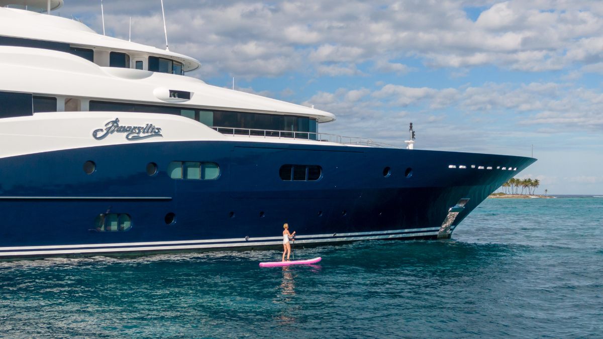 A person stands on a pink paddleboard in calm waters next to a large luxury yacht named "Anastasia." The sky is partly cloudy with a few palm trees visible in the background.