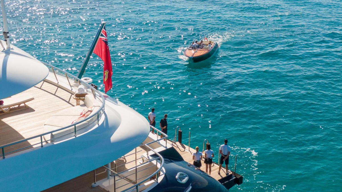 A luxurious yacht is anchored in vibrant blue waters, with crew members standing on the deck. A small wooden boat approaches the yacht, carrying passengers. A flag is flying at the back of the yacht. Sunlight sparkles on the water's surface.