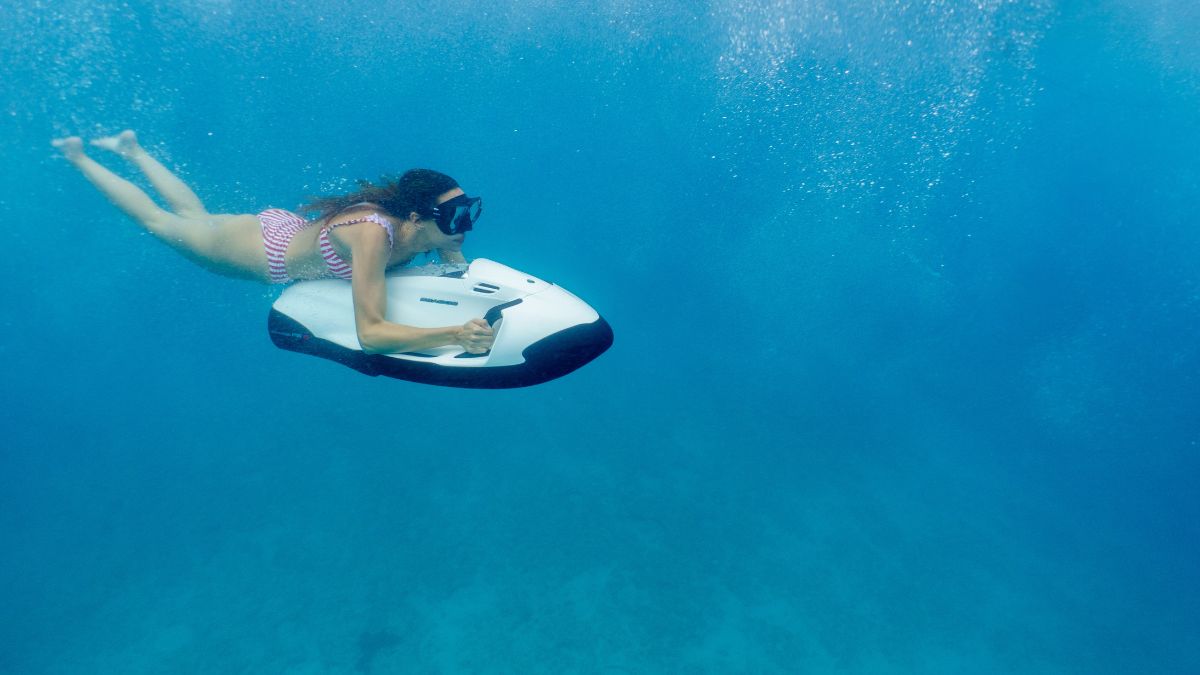 A swimmer in a striped swimsuit glides underwater on a white snorkeling scooter, surrounded by clear blue water and bubbles.