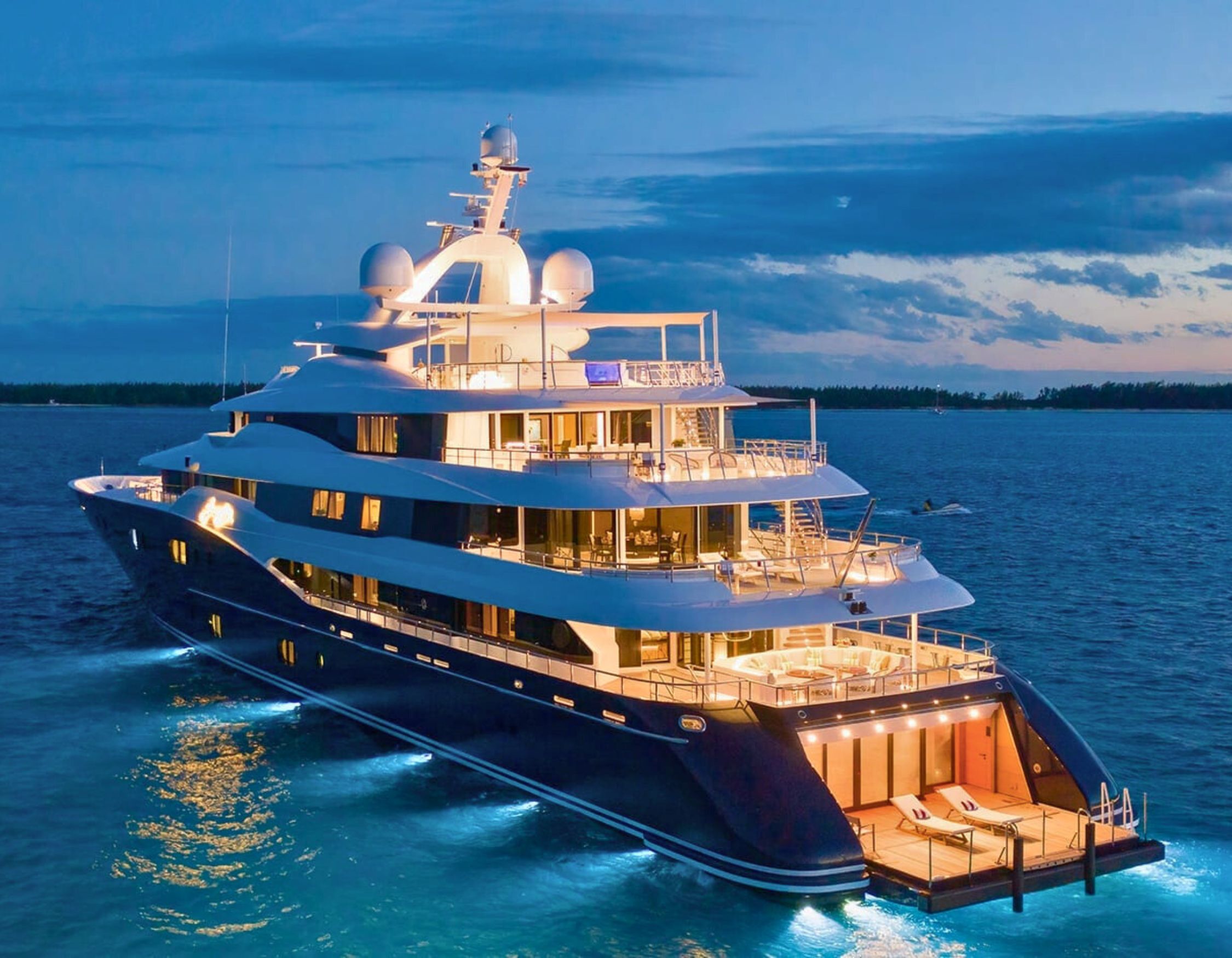 A luxurious yacht docked in calm waters during twilight, illuminated by warm lights. The multi-deck vessel features sleek lines and modern design, with a spacious upper deck and large windows reflecting the evening sky.
