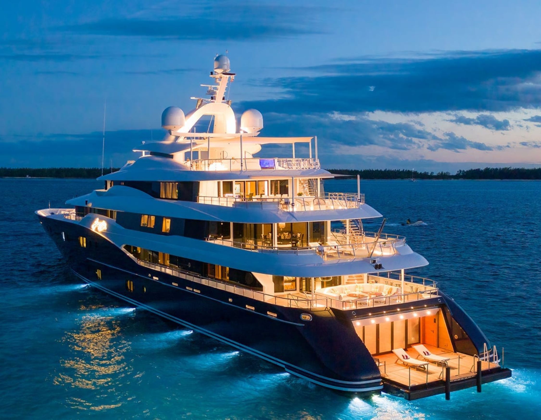 A luxurious yacht illuminated at night, anchored in clear blue water with a stunning sunset in the background. The yacht features multiple decks, large windows, and sleek lines, creating an elegant and opulent appearance.