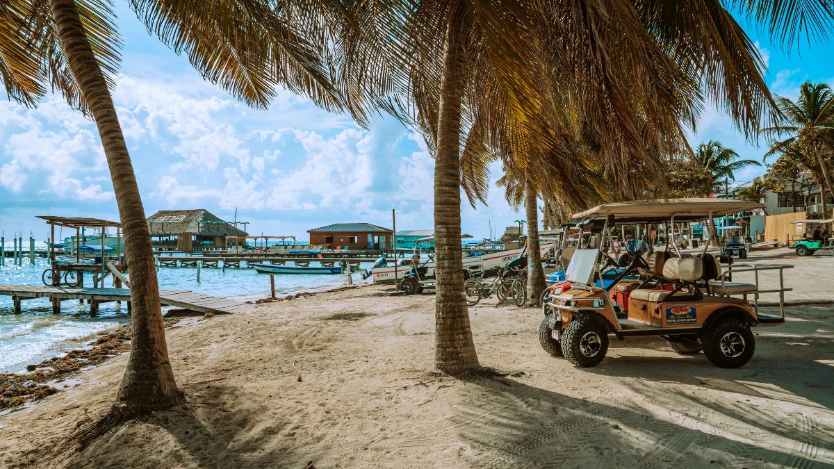 A golf cart parked on a sandy beach surrounded by palm trees, with boats moored in the water and rustic wooden structures in the background under a partly cloudy sky.