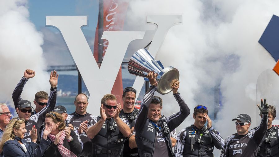 A victorious sailing team celebrates on the podium, holding a large trophy while surrounded by cheering supporters and billowing smoke. The backdrop features a prominent "LV" logo.