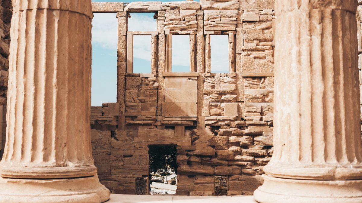 Ancient stone columns frame a view of a ruined building with gaps where windows once existed, showcasing weathered stone walls against a blue sky.