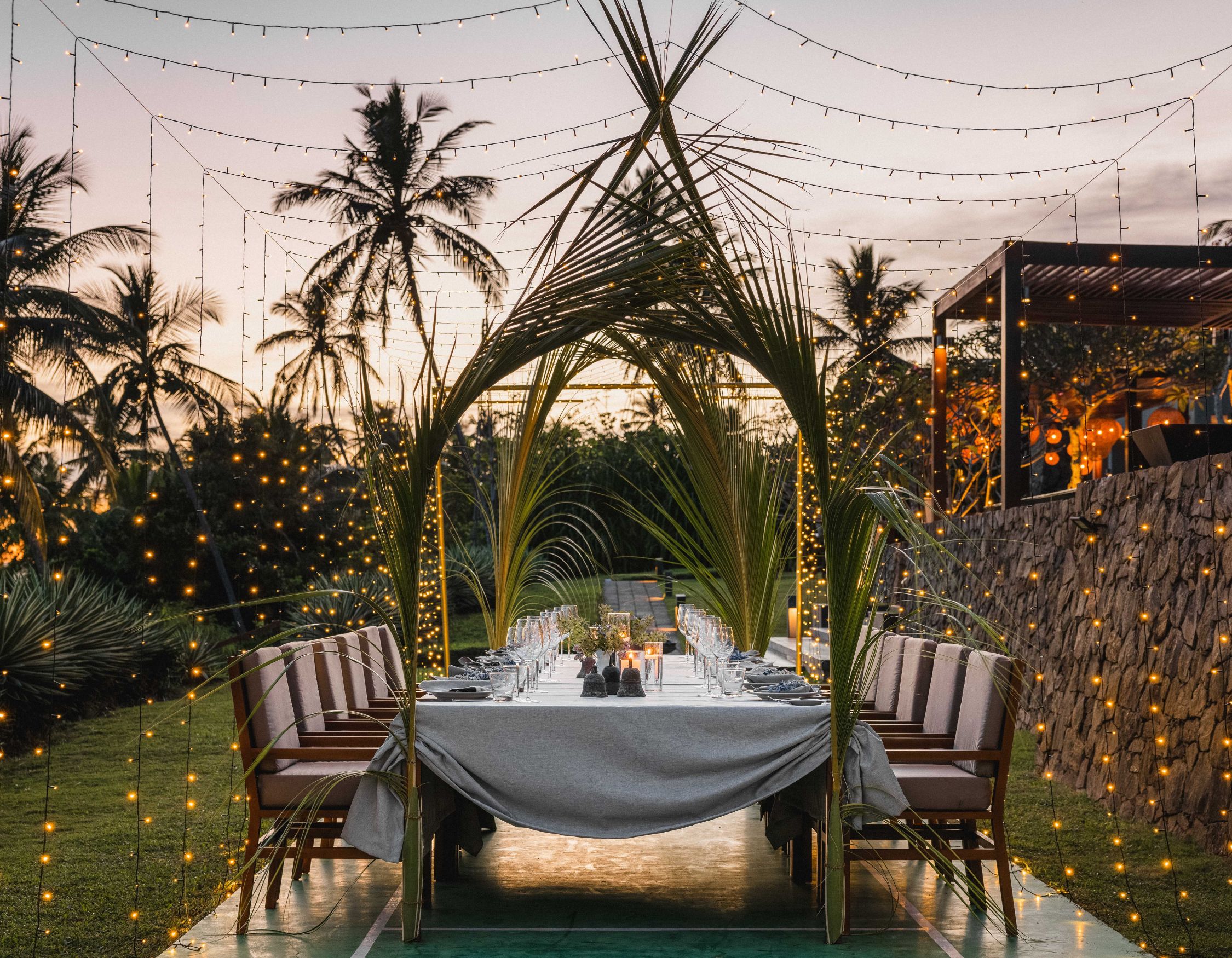 An elegantly arranged outdoor dining setup surrounded by palm trees, decorated with string lights. The table is set with glassware and tableware, with a sunset backdrop creating a warm, inviting atmosphere.