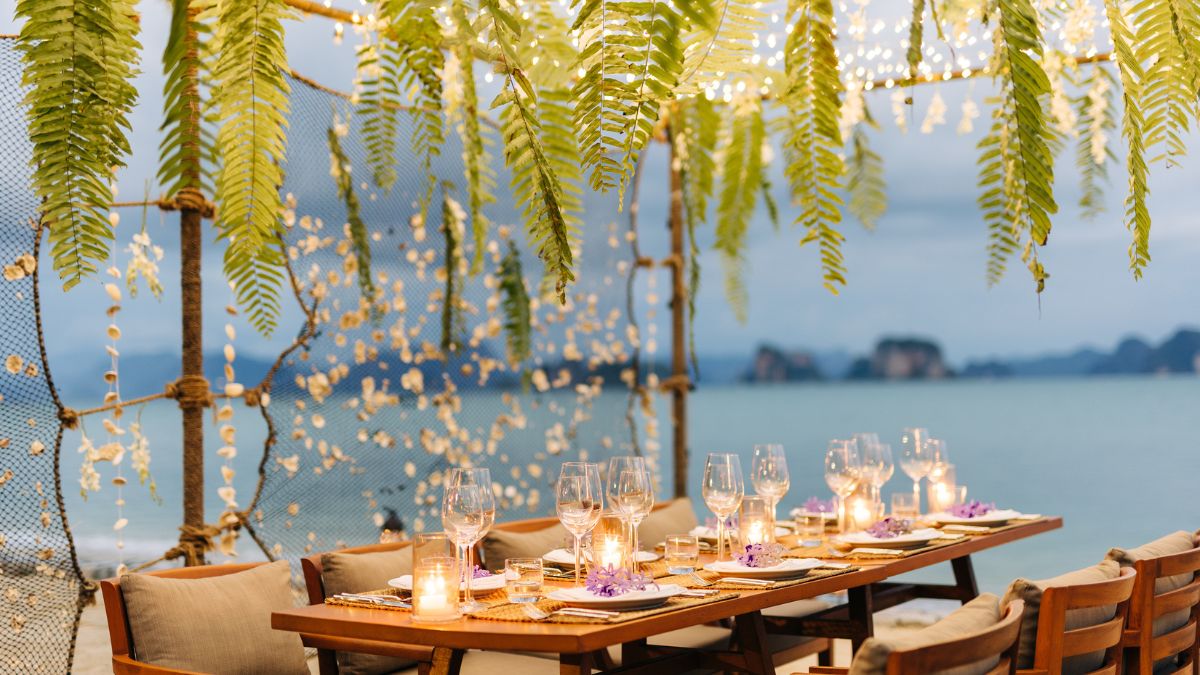A beautifully arranged outdoor dining table set for a special occasion. The table features elegant glassware and floral decorations, surrounded by hanging ferns and twinkling fairy lights, with a scenic view of the water and distant islands in the background.