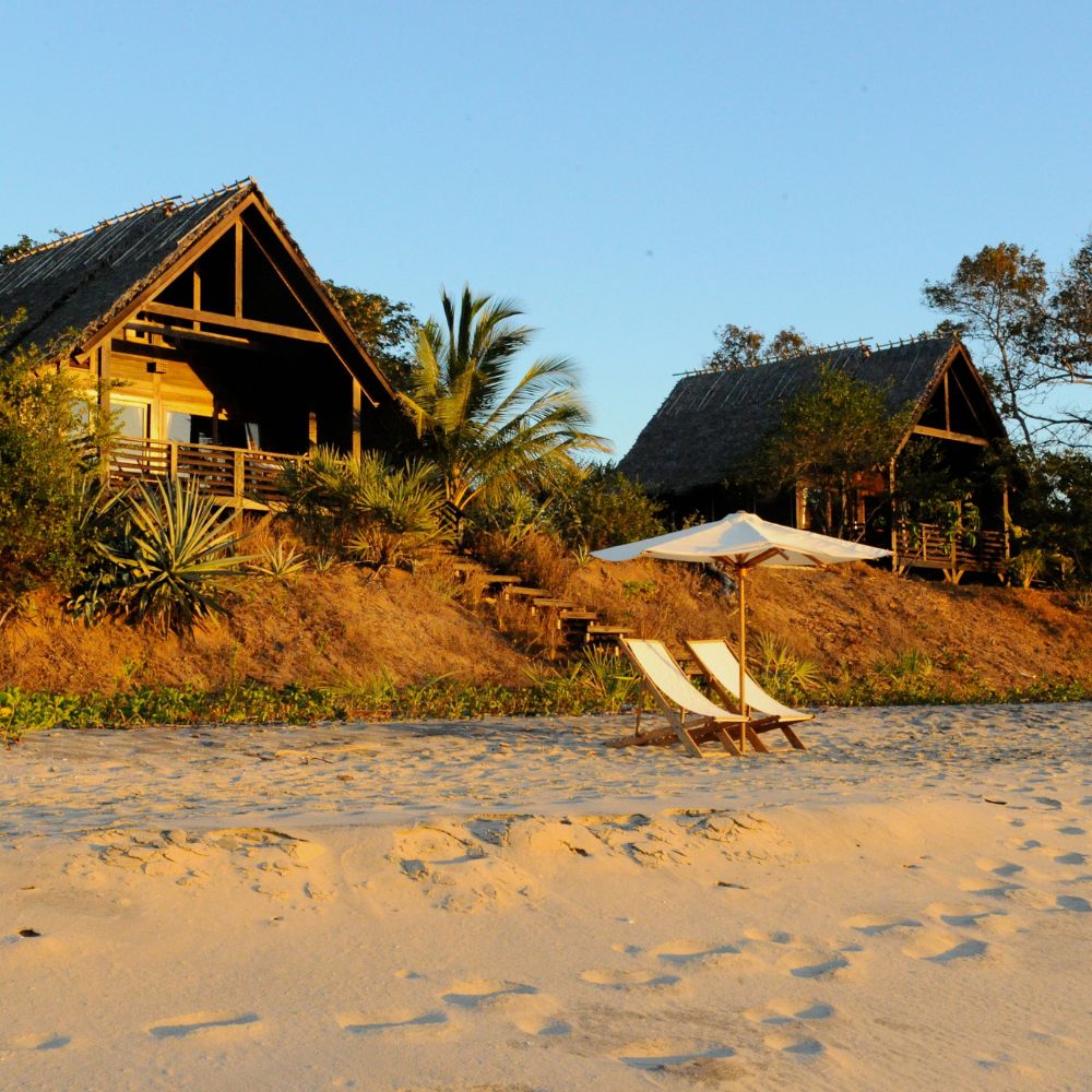 Two wooden beach huts surrounded by greenery, with white sun loungers and an umbrella on sandy shores under a clear blue sky.
