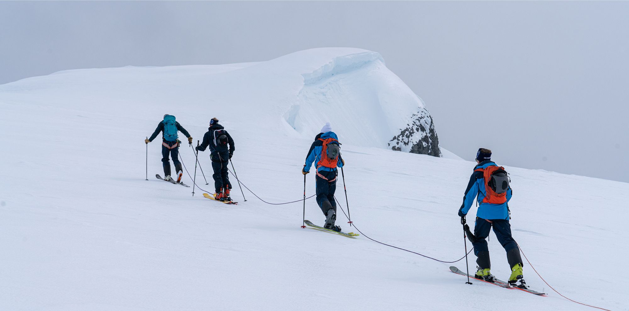 Four climbers ascend a snowy slope, tethered together with ropes. They wear winter gear and skis, with a rocky cliff visible in the background. The landscape is overcast and shrouded in mist.