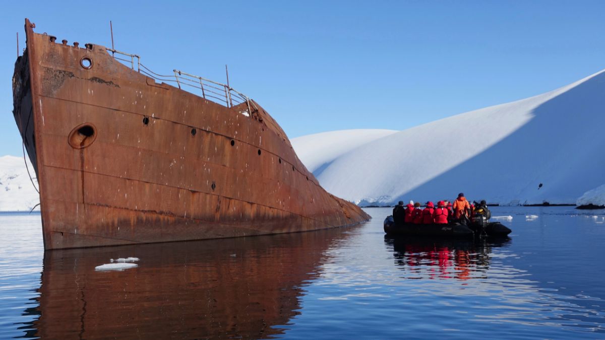 A rusted shipwreck partially submerged in calm waters, with a group of people in a small boat nearby. Snow-covered mountains rise in the background under a clear blue sky.