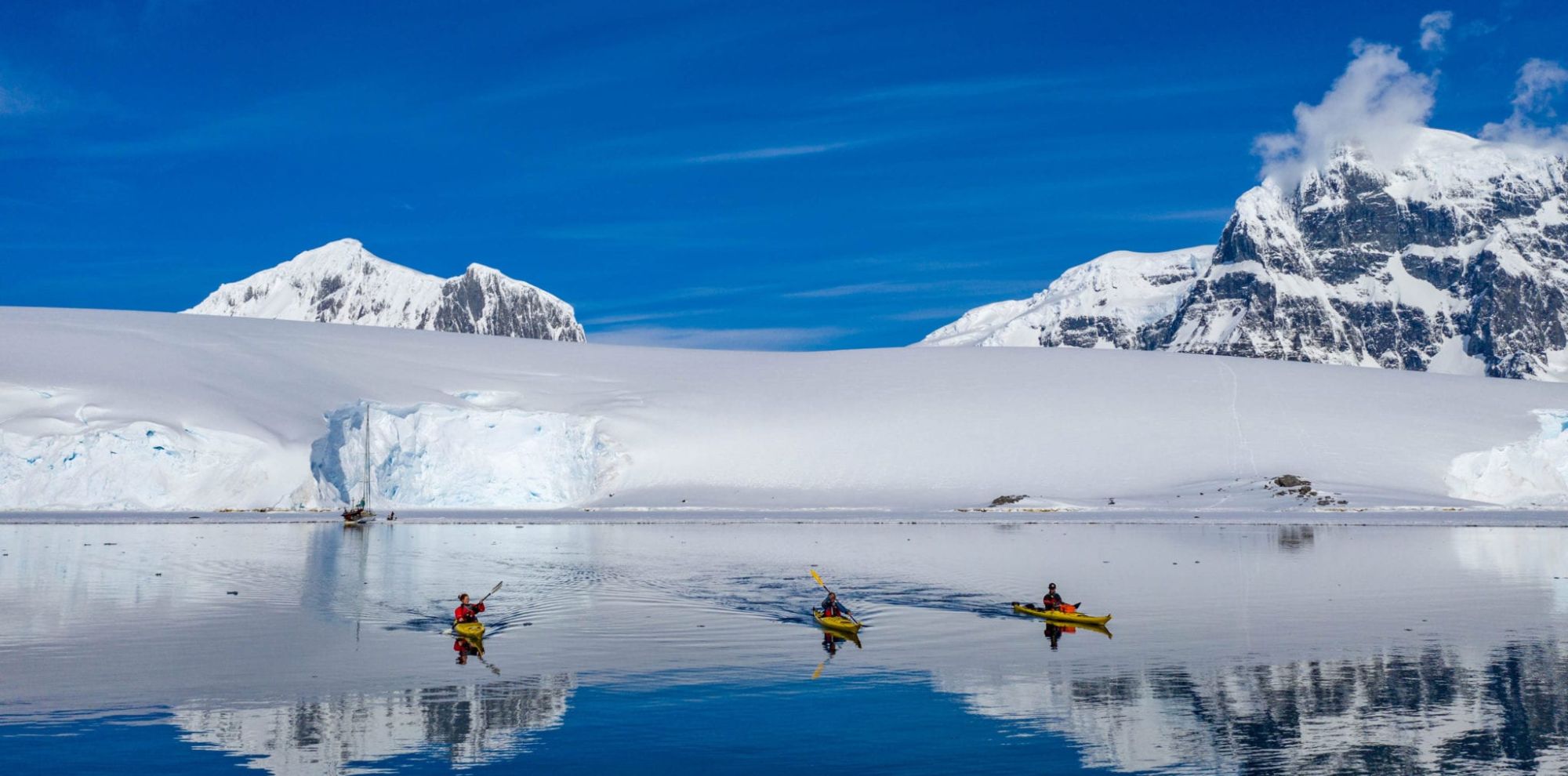 Three kayakers paddle through calm, reflective waters with majestic snow-covered mountains and icebergs in the background. Clear blue skies enhance the serene Arctic landscape.