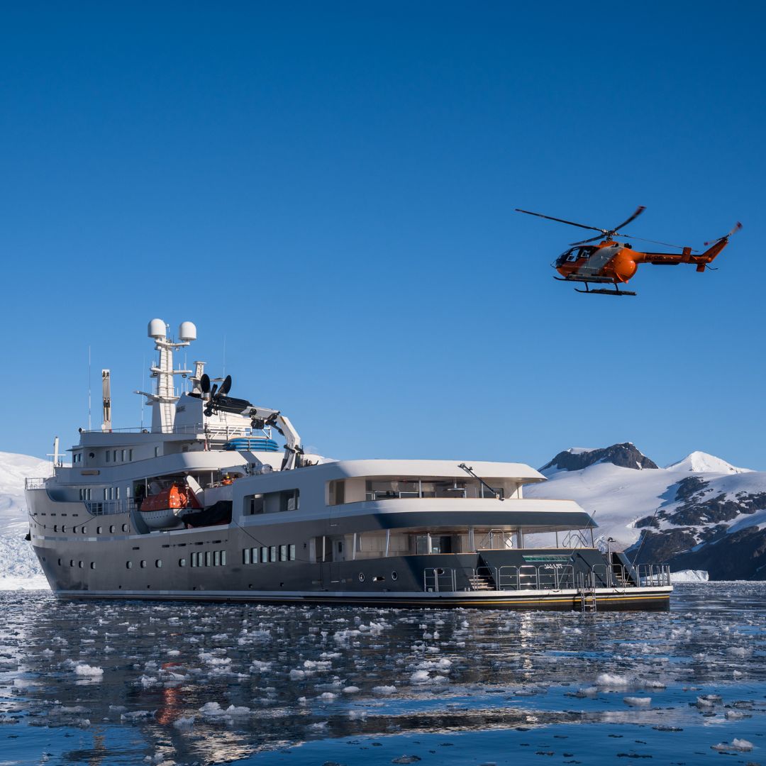 A large expedition ship navigates through icy waters filled with small ice chunks, with an orange helicopter flying nearby.