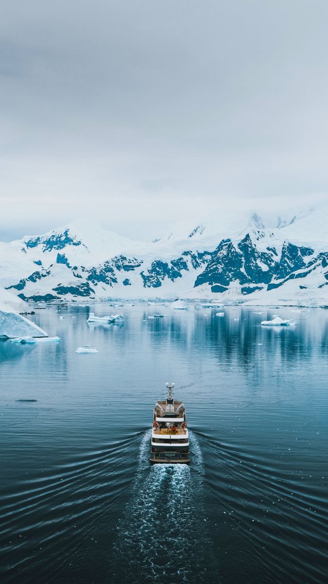 Yacht navigates through calm, icy waters surrounded by towering mountains and floating icebergs. The scene is muted with overcast skies, creating a tranquil atmosphere.