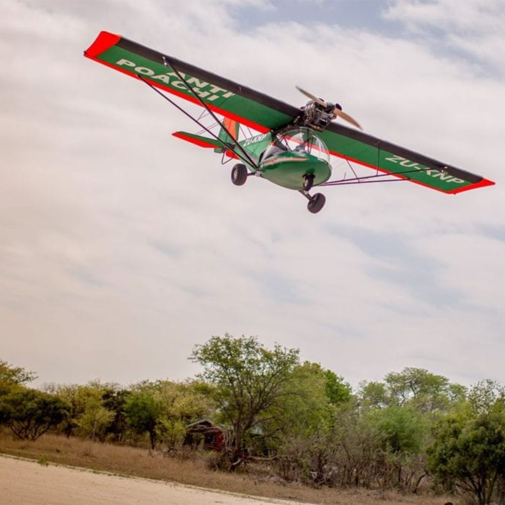 A green and red aircraft flies low above a sandy area, with "Anti Poach" written prominently on its wings. In the background, trees and a small building are visible under a cloudy sky.