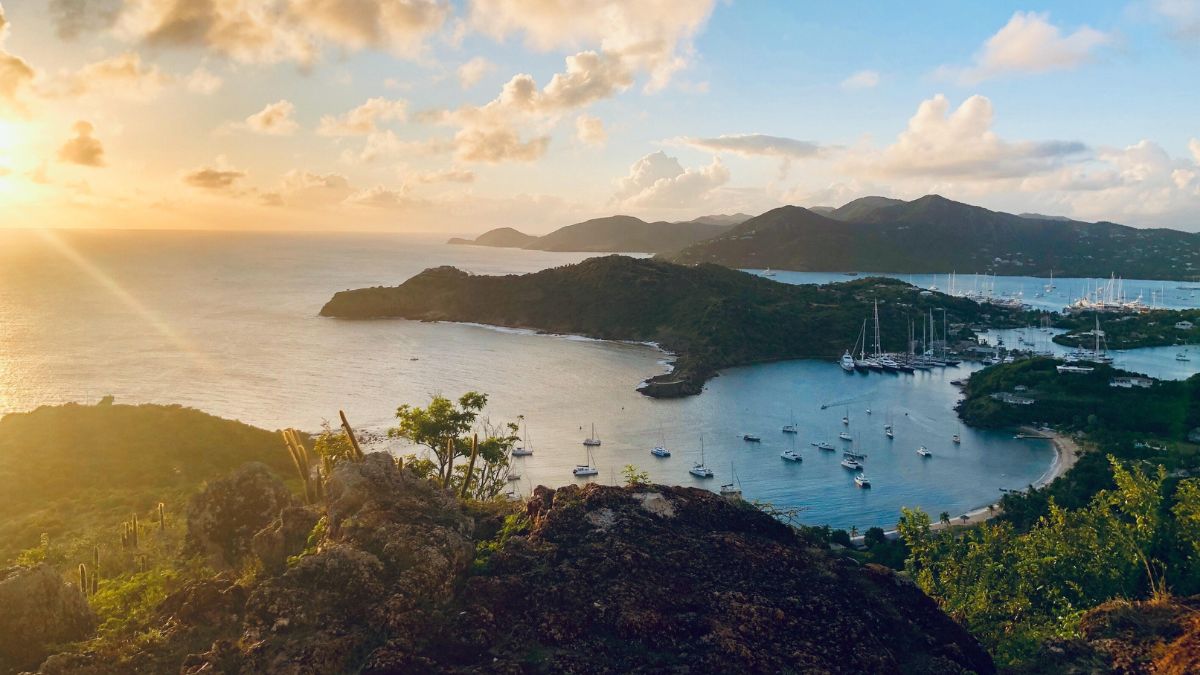 A picturesque view of a serene bay at sunset, featuring calm waters dotted with sailboats. Lush green hills surround the coastline, with distant mountains under a partly cloudy sky illuminated by warm sunlight.